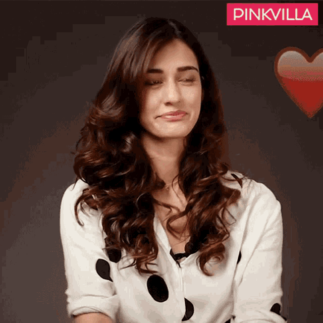 a woman wearing a white shirt with black polka dots is standing in front of a pinkvilla sign