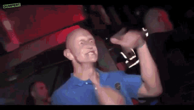 a bald man in a blue shirt is making a funny face while standing in a dark room .