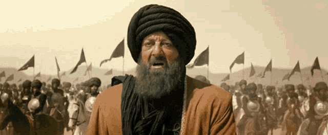 a man with a beard wearing a turban stands in front of a crowd of soldiers on horses .