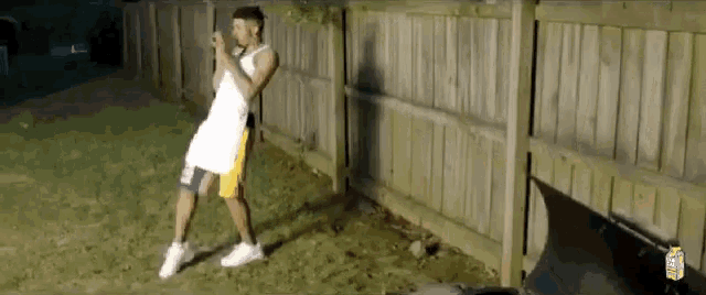 a man in a white tank top and yellow shorts is standing in front of a wooden fence
