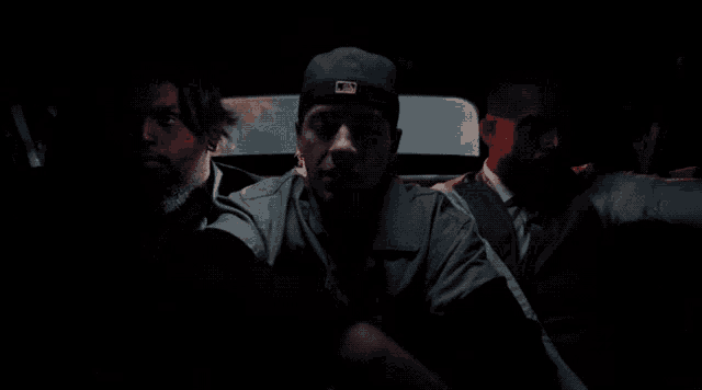 a man wearing a new era hat sits in the back seat of a car with two other men