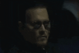 a man wearing sunglasses and a black shirt is looking at the camera in a dark room .