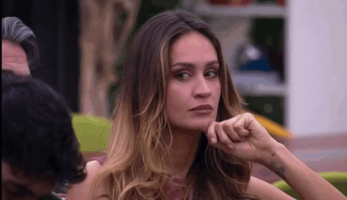 a woman with long hair is sitting at a table with her hand on her chin and looking at the camera .