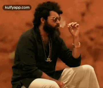 a man with a beard is smoking a cigarette while wearing sunglasses .