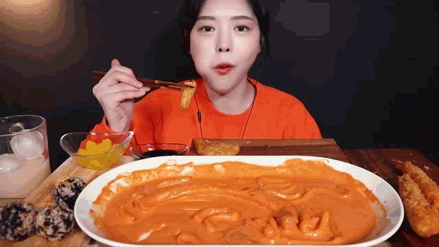 a woman in an orange sweater is eating a plate of food with chopsticks