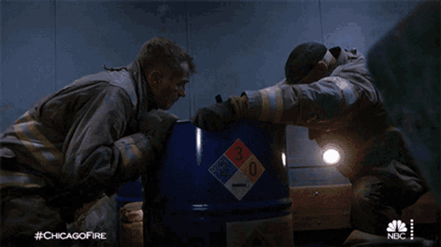 two firefighters are standing next to a barrel with a nbc logo in the background