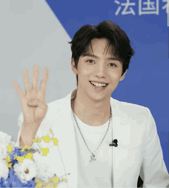 a man wearing a white jacket and a necklace is smiling and giving the number four