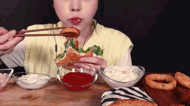 a woman is eating a sandwich with chopsticks next to a bowl of ketchup