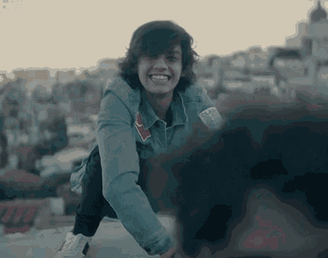 a young man in a denim jacket is smiling while kneeling down on a ledge .