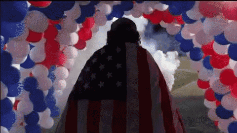 a man in an american flag cape is standing in front of a bunch of red , white and blue balloons .