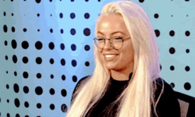 a blonde woman wearing glasses and a black shirt is smiling in front of a blue background with polka dots .
