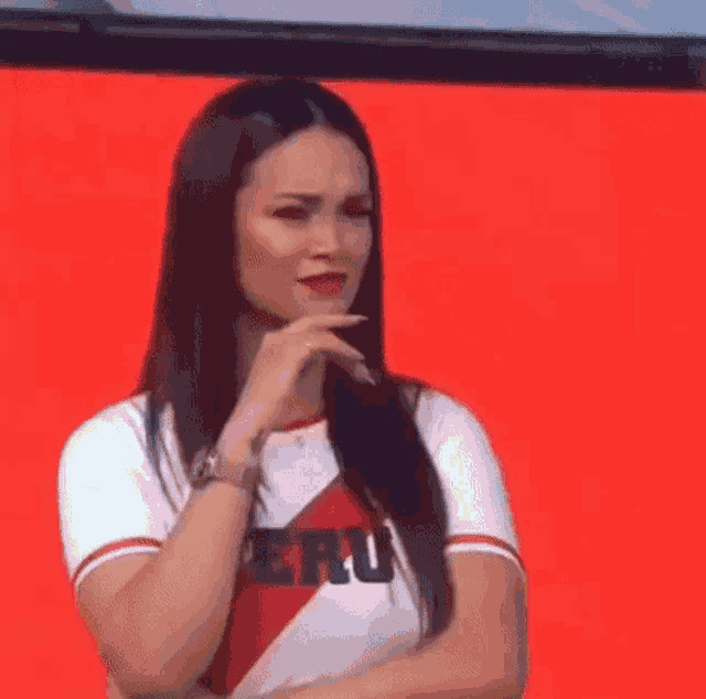 a woman wearing a peru shirt is standing with her arms crossed and her hand on her chin .