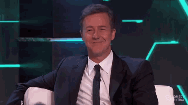 a man in a suit and tie is sitting on a couch and covering his mouth with his hand .