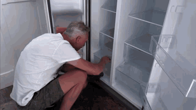 a man in a white shirt is kneeling in front of an empty fridge