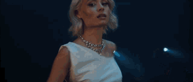 a woman in a white dress and necklace is standing in front of a blue background .