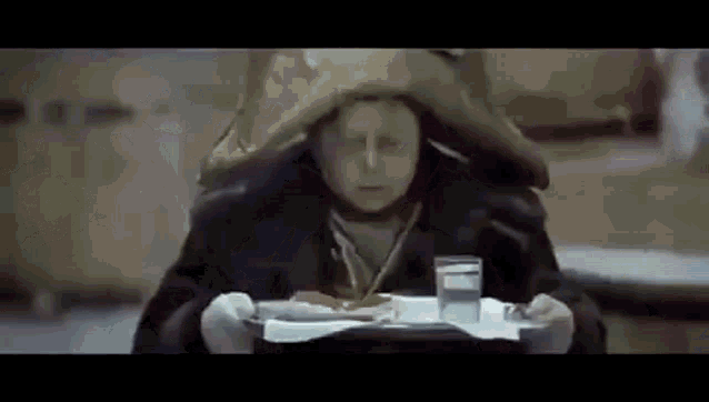 an elderly woman is sitting at a table holding a tray of food and a glass of water .