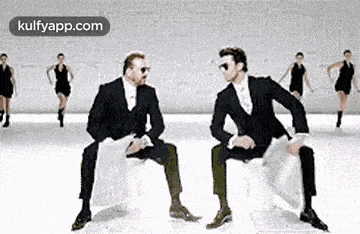 two men in suits are sitting on chairs in front of a group of women dancing .