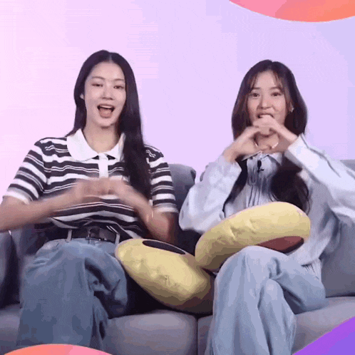two women are sitting on a couch with pillows and making a heart shape with their hands