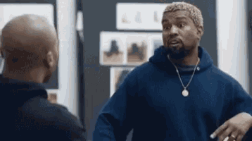 a man in a blue hoodie and necklace is talking to another man in a room .