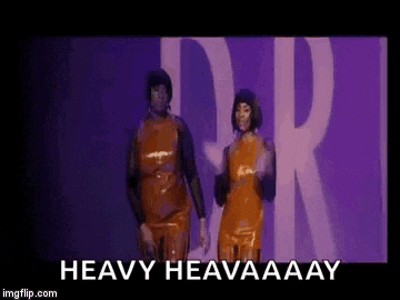 two women in orange latex dresses are dancing on a stage with the words `` heavy heaaaay '' above them .