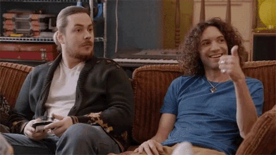 two men are sitting on a couch playing a video game and one of them is giving a thumbs up