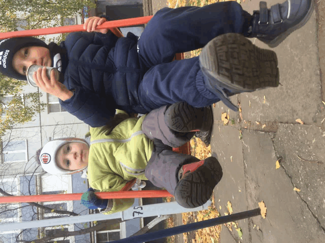 a boy wearing an adidas hat sits on a swing with another child