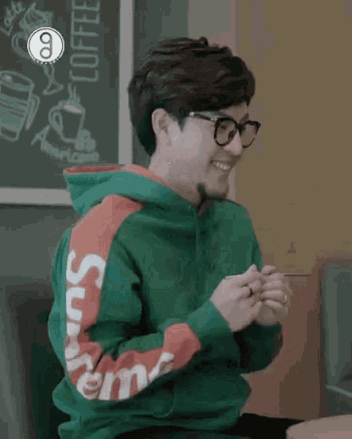 a man wearing glasses and a green hoodie is sitting at a table and smiling .