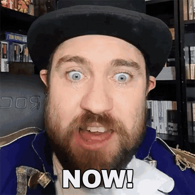 a man with a beard wearing a hat and a blue jacket says now