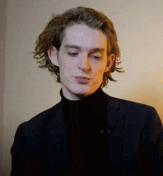 a young man wearing a black turtleneck and a suit looks at the camera