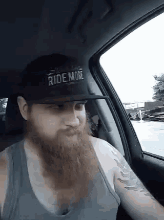 a man with a beard wearing a hat that says ride more is sitting in a car .