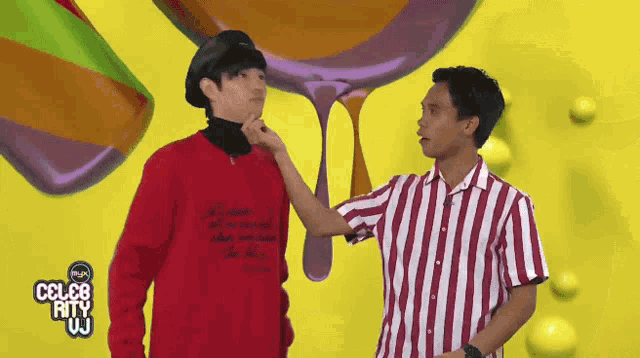 a man in a striped shirt is touching another man 's face in front of a yellow background that says celeb rity