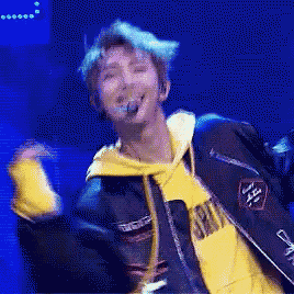 a man with blue hair is wearing a yellow hoodie and a black jacket while dancing on a stage .