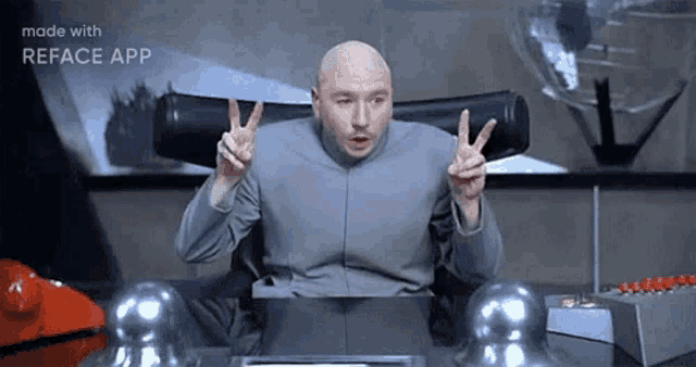 a bald man is sitting at a desk making a peace sign .
