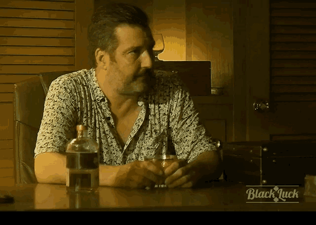 a man sits at a table with a bottle of whiskey and a glass of whiskey in front of a black luck logo