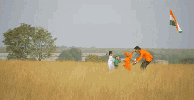 a man in an orange shirt talks to two children in a field with a flag in the background