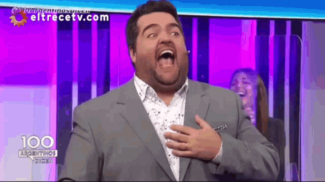 a man in a suit is laughing with his hands on his chest while a woman laughs behind him .