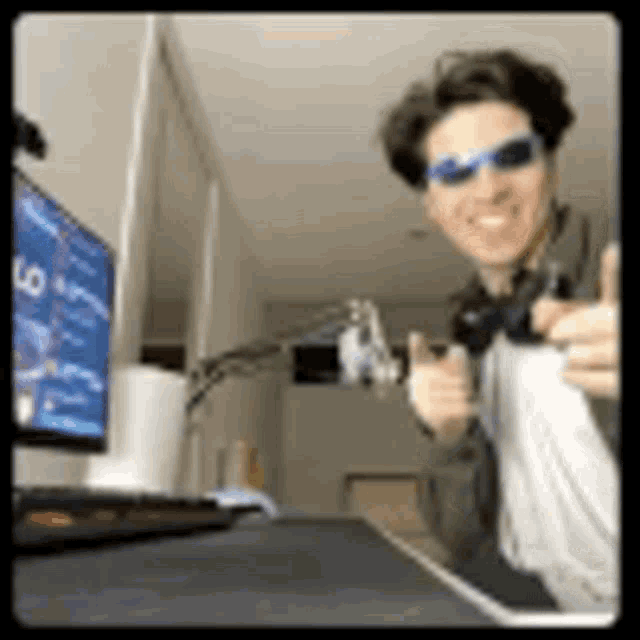 a man wearing sunglasses and headphones is giving a thumbs up in front of a computer .
