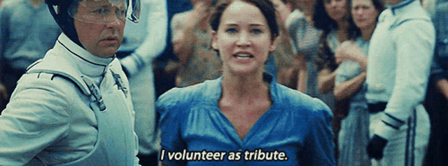 a woman in a blue shirt says " i volunteer as tribute " in front of a crowd of people