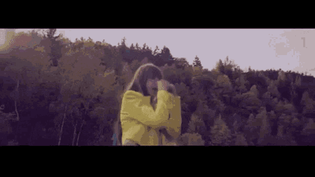 a woman in a yellow jacket is standing in front of a forest and taking a picture .
