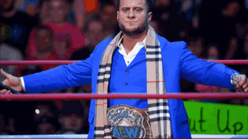 a wrestler in a blue suit and scarf stands in a ring with a sign that says shut up
