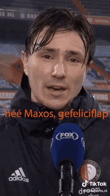 a man wearing an adidas jacket stands in front of a blue fox sports microphone