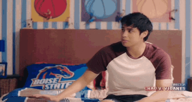 a young man sits on a bed with a boise state pillow on it
