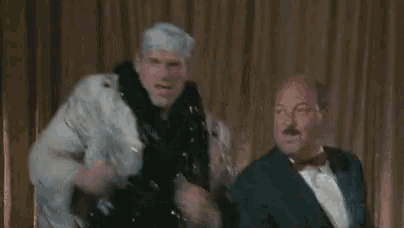 a man in a bandana is singing into a microphone while another man in a tuxedo looks on .