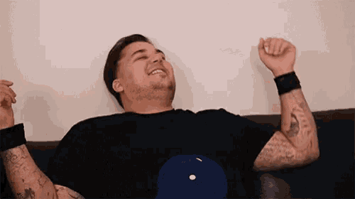 a man with tattoos is sitting on a couch with his arms in the air and laughing .