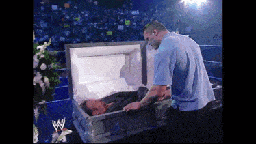 a man in a blue shirt stands next to a coffin with the letter w on it