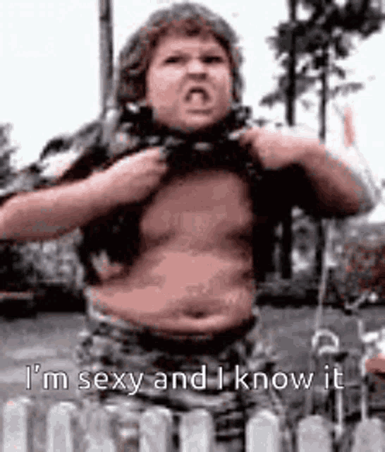 a fat boy is standing in front of a fence and taking off his shirt while making a funny face .