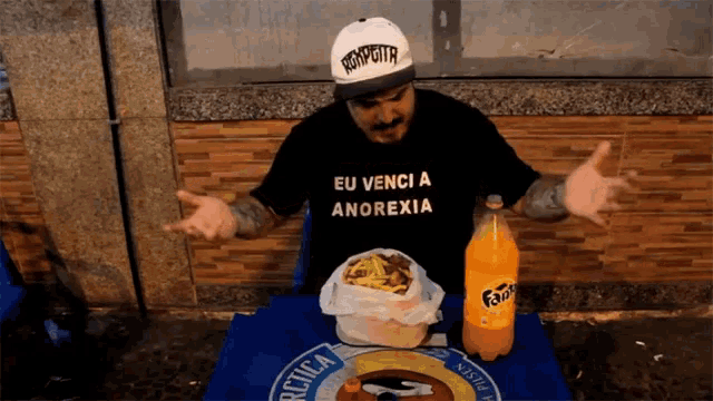 a man wearing a shirt that says eu venci a anorexia sits at a table with a bottle of fanta