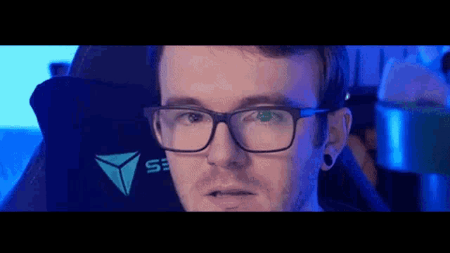 a man wearing glasses is sitting in a gaming chair looking at the camera .