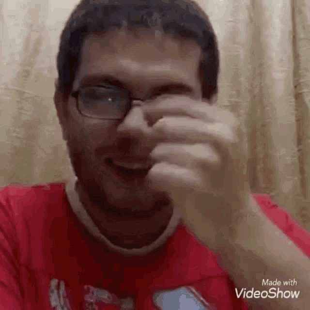 a man wearing glasses and a red shirt is smiling and covering his face with his hands .