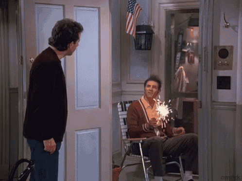 a man in a brown sweater is standing next to a man in a chair holding a sparkler